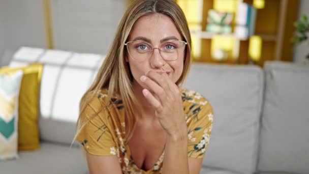 jong blond vrouw zitten op bank doen dicht mond gebaar thuis - Video