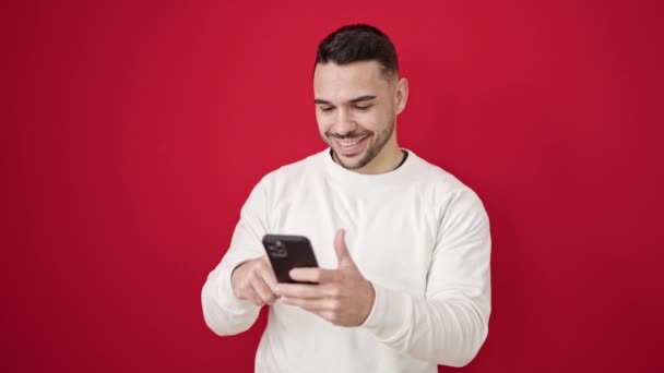 Молодий іспаномовний чоловік використовує смартфон, посміхаючись над ізольованим червоним тлом
 - Кадри, відео