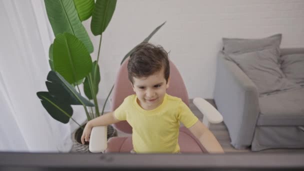 Feliz niño sentado en la silla frente a la computadora de escritorio portátil viendo dibujos animados, preparándose para aprender en línea lección de educación a distancia para los niños. Niño divirtiéndose en casa - Imágenes, Vídeo