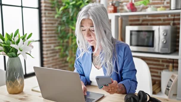 Orta yaşlı, gri saçlı, dizüstü bilgisayar ve akıllı telefon kullanan bir kadın yemek odasındaki masada oturuyor. - Video, Çekim