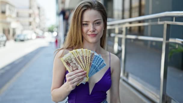 Joven mujer rubia sonriendo confiado sosteniendo hungary forint billetes en la calle - Imágenes, Vídeo
