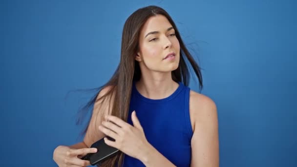 Jeune belle femme hispanique souriant cheveux peignants confiants sur fond bleu isolé - Séquence, vidéo