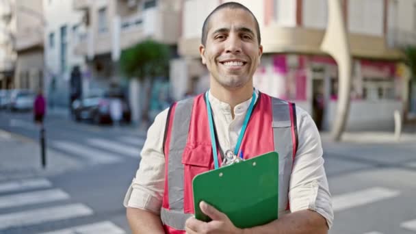 Giovane uomo ispanico volontario sorridente tenendo appunti in strada - Filmati, video