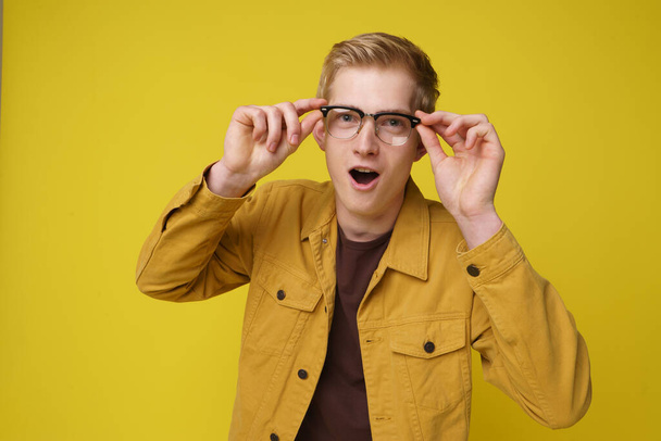 Здивований молодий 20-річний чоловік з відкритим ротом торкається окулярів двома руками. Захоплений блондинка молодий чоловік ізольований жовтий фон - Фото, зображення