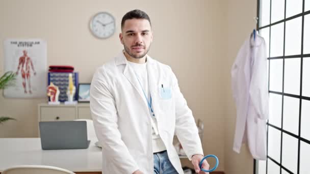 Νεαρός Ισπανός γιατρός χαμογελά σίγουρος κρατώντας στηθοσκόπιο στην κλινική - Πλάνα, βίντεο