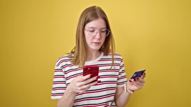 Mladé blondýny žena nakupování s smartphone a kreditní karty hledá rozrušený přes izolované žluté pozadí - Záběry, video