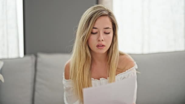 Joven mujer rubia leyendo documento sentado en el sofá en casa - Metraje, vídeo
