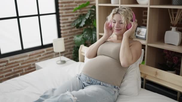 Młoda ciężarna kobieta słuchająca muzyki zakładająca słuchawki na brzuch w sypialni - Materiał filmowy, wideo