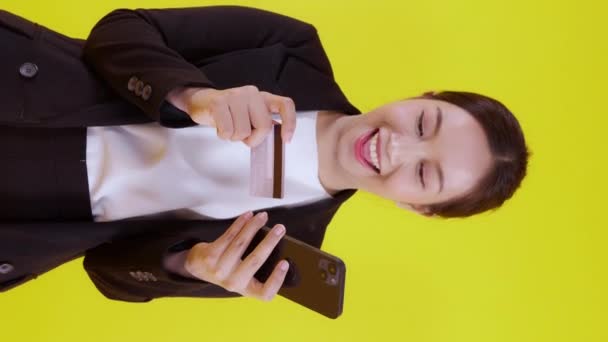 Portret jonge Aziatische zakenvrouw houden credit card en op zoek naar smart phone voor winkelen online geïsoleerde gele achtergrond, zakenvrouw op zoek debetkaart en smartphone, financieel concept. - Video