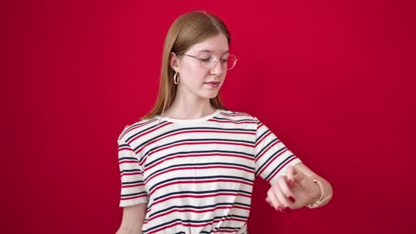 Young blonde woman looking watch with unhappy expression over isolated red background - Footage, Video