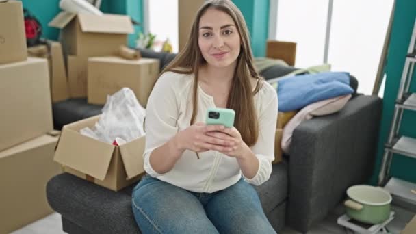 Mladý krásný hispánec žena pomocí smartphone sedí na pohovce v novém domově - Záběry, video