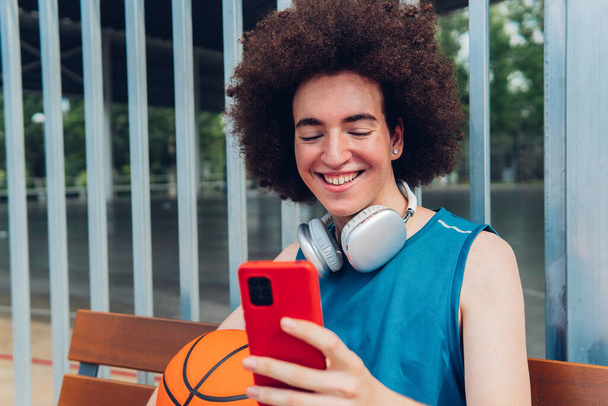 Αγόρι που χρησιμοποιεί το τηλέφωνο κάθεται σε ένα παγκάκι ενός γηπέδου μπάσκετ - Φωτογραφία, εικόνα