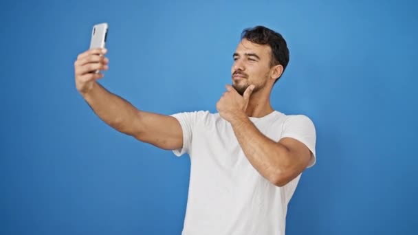 孤立した青い背景の上にスマートフォンでセルフィーを作る自信を持って微笑む若いヒスパニック男性 - 映像、動画