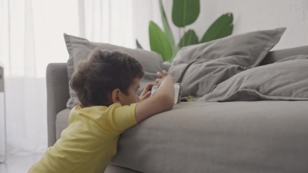 Симпатичный малыш играет со смартфоном, смотрит мультики, трогает мобильный экран, сидит на полу в гостиной один дома  - Кадры, видео