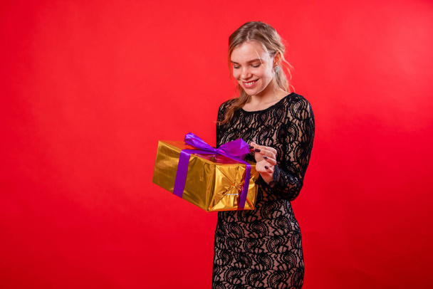 Portrait of an attractive young woman in a black dress with a gift in her hands, isolated on a red background. - Fotografie, Obrázek