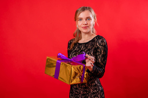 Portrait of an attractive young woman in a black dress with a gift in her hands, isolated on a red background. - Zdjęcie, obraz