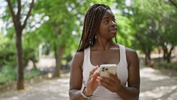 Αφροαμερικανή γυναίκα που χρησιμοποιεί smartphone χαμογελώντας στο πάρκο - Πλάνα, βίντεο