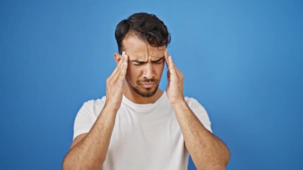 Joven hombre hispano estresado de pie sufriendo por dolor de cabeza sobre fondo azul aislado - Metraje, vídeo
