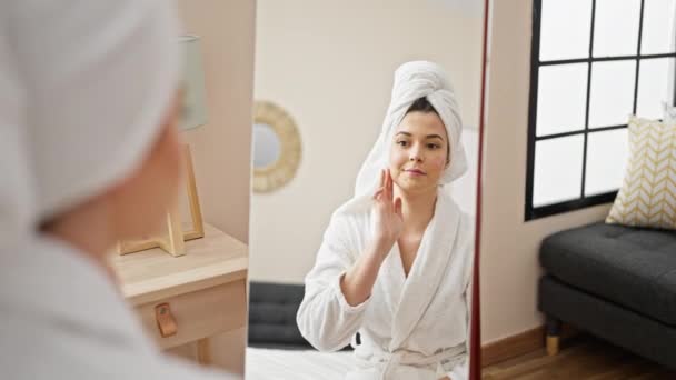 Jovem bela mulher hispânica vestindo roupão de banho tocando rosto olhando no espelho no quarto - Filmagem, Vídeo