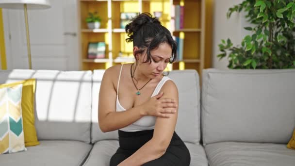 Young beautiful hispanic woman scratching arm sitting on sofa at home - 映像、動画