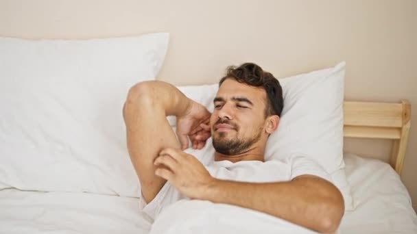 Joven hispano acostado en la cama arañando el brazo en el dormitorio - Imágenes, Vídeo