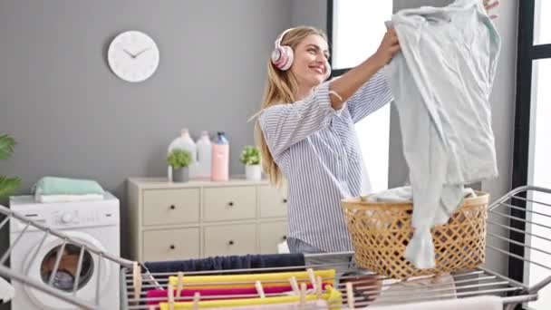 Fiatal szőke nő zenét hallgat, ruhákat lóg a ruhaszárítón a mosodában. - Felvétel, videó