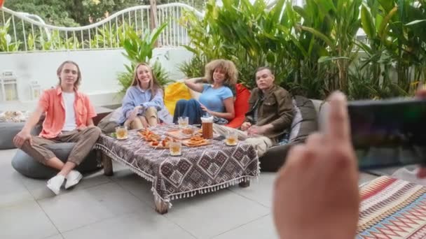 Рука делает групповое фото друзей на смартфоне, пока они тусуются вместе в кафе на открытом воздухе - Кадры, видео