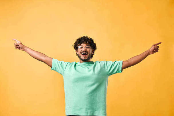 Handsome young emotional curly-haired man in a turquoise t-shirt on a yellow background shows his hands to the side and laughs. Emotion of joy, happiness. Advertising,copy space.                                - Фото, изображение