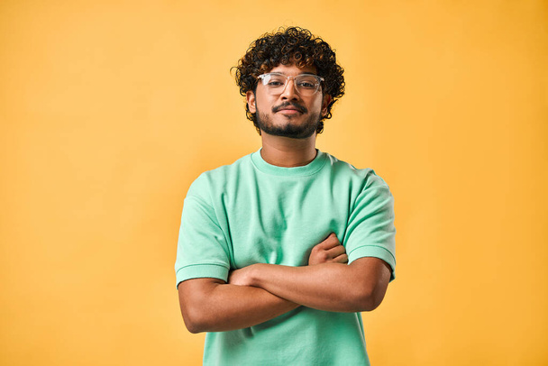 Portrait of a handsome young man in glasses and a turquoise t-shirt with his arms crossed looking at the camera.                                - Foto, imagen