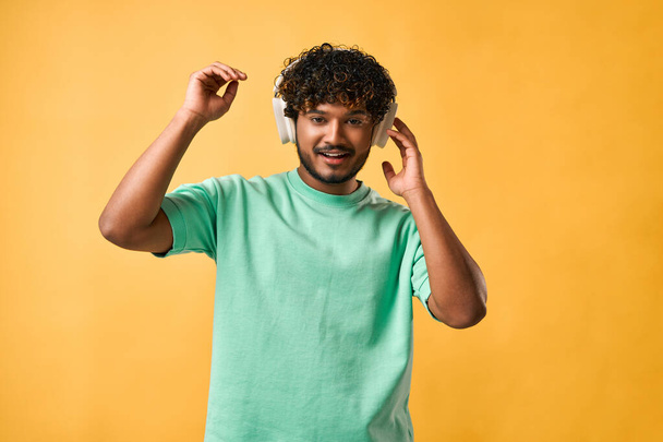 Portrait of a curly-haired Indian man in a turquoise t-shirt adjusting wireless headphones and looking at the camera.                                - 写真・画像