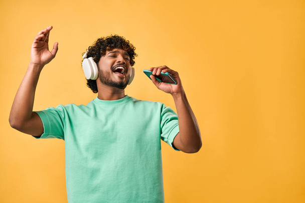 Portrait of handsome curly Indian man in turquoise t-shirt and singing with phone in wireless headphones enjoying music and gesturing emotionally. - Photo, Image
