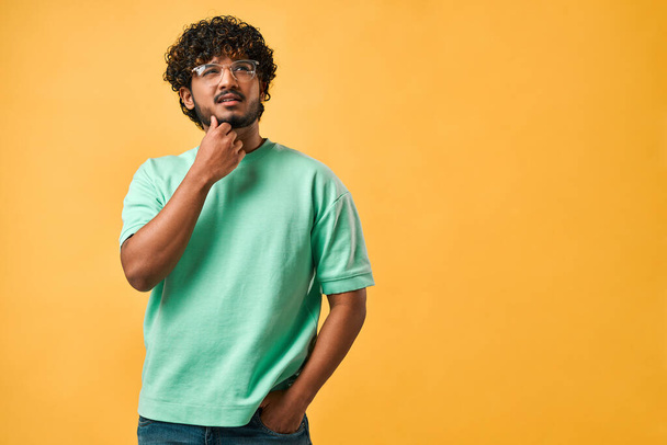 Portrait of a handsome curly-haired Indian man in a turquoise t-shirt and glasses thoughtfully scratching his chin and looking up. The emotion of thoughtfulness, doubt. Copy space. - Фото, зображення