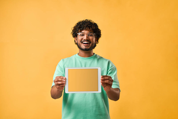 Portrait of a handsome curly-haired Indian man in a turquoise t-shirt and glasses thoughtfully scratching his chin and looking up. The emotion of thoughtfulness, doubt. Copy space. - 写真・画像