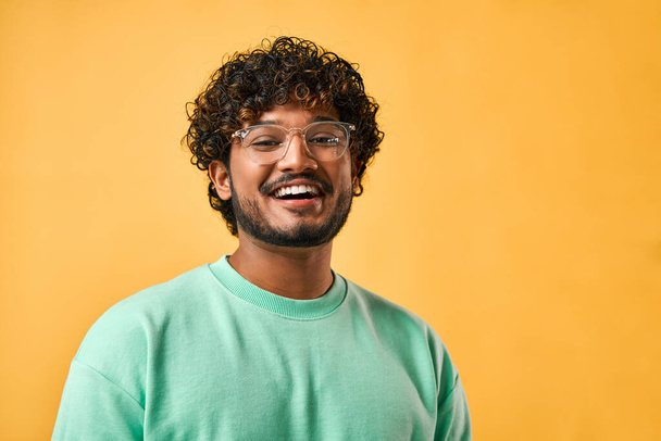 Retrato de cerca de un guapo indio de pelo rizado con una camiseta turquesa y gafas riendo y mirando a la cámara. - Foto, imagen