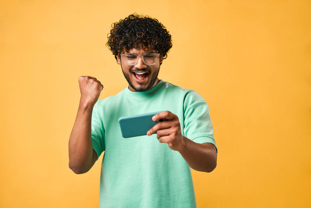 Portrait of a handsome Indian man with curly hair in a turquoise t-shirt and glasses standing on a yellow background and playing on the phone. Emotion of joy, celebration of victory.                                - 写真・画像