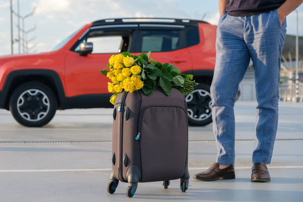 Cropped view of a man with luggage and a bouquet of flowers in the parking lot. Travel, vacation concept. - Φωτογραφία, εικόνα