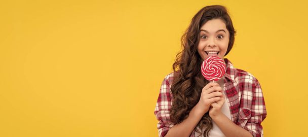 happy kid with long curly hair in checkered shirt lick lollipop caramel candy on yellow background, childhood. - Fotografie, Obrázek