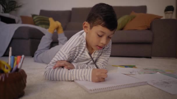 Λατινικά μικρό αγόρι επικεντρώνεται σε σχέδια με το χρωματισμό στο σημειωματάριο στο πάτωμα του σπιτιού. Παιδιά ξαπλωμένα στο σαλόνι ζωγραφίζοντας χαλιά στον ελεύθερο χρόνο. Έννοια της homeschooling και την ανάπτυξη των παιδιών. - Πλάνα, βίντεο