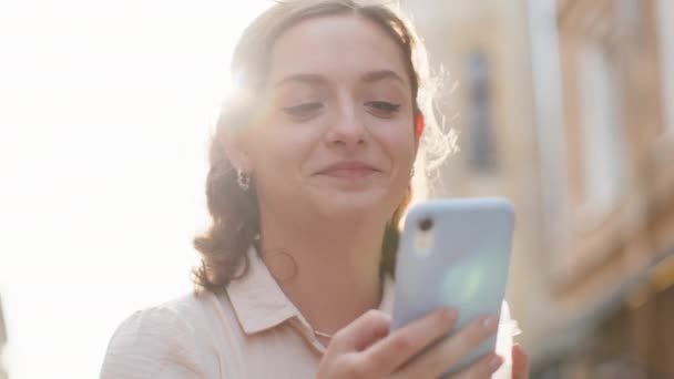 スマートフォンの入力テキスト回答メッセージを使用してかなり若い女性を笑顔モバイル画面のソーシャルメディアアプリを見てオンラインチャット。女の子の観光客は氷と冷たいコーヒーを飲む街の通りのベンチに座っています - 映像、動画