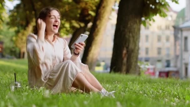 Jeune femme utilisent smartphone mobile célébrant gagner de bonnes nouvelles de message, victoire de jackpot de loterie, cadeau en ligne. Fille heureuse assise sur l'herbe dans le parc urbain de la ville coucher de soleil en plein air fond. Mode de vie - Séquence, vidéo