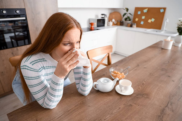 Allergic young woman sneezing into tissue in kitchen - Zdjęcie, obraz