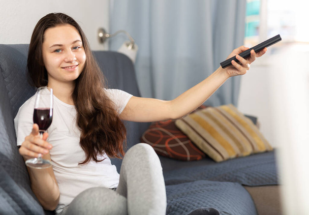 Portrait of young woman with glass of wine sitting on sofa at home watching movie - Photo, Image