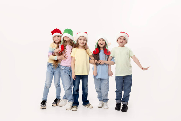 С Новым годом и Рождеством! Группа веселых счастливых детей в праздничном Санта-Клаусе и эльфийских шляпах на белом фоне студии. Дети обнимаются и веселятся - Фото, изображение