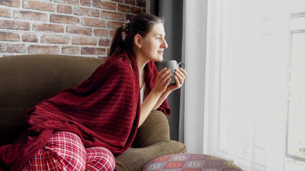 Beautiful woman in pajamas drinking tea on the sofa at morning. Female resting at home, lifestyle and relaxation - Foto, Imagem