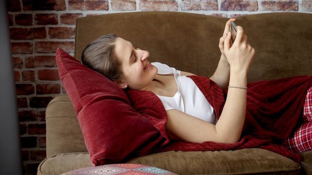 Joven mujer sonriente descansando en el sofá en la sala de estar y utilizando el teléfono inteligente. Personas relajadas en casa, comunicaciones, trabajo remoto - Foto, Imagen