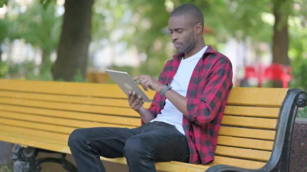 Giovane uomo africano utilizzando tavoletta mentre seduto all'aperto su una panchina - Filmati, video