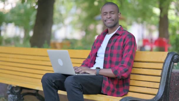 Młody Afrykanin uśmiecha się do kamery podczas pracy na zewnątrz laptopa - Materiał filmowy, wideo