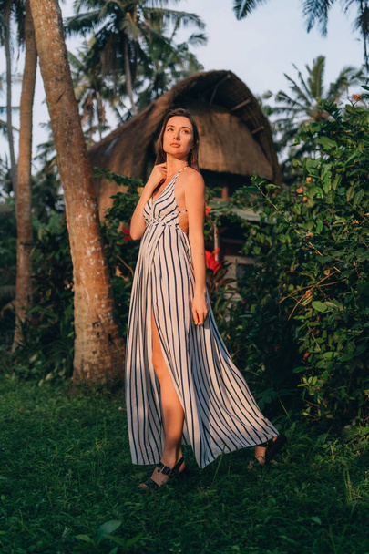 Portrait of beautiful lady in white striped dress with balinese vila background. Young model girl posing in tropical jungle nature - Fotó, kép