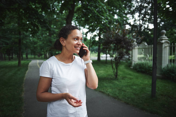Piękna, wieloetniczna młoda kobieta w białej koszulce, uśmiechnięta, rozmawiająca na smartfonie podczas spaceru uliczką parku miejskiego w letni dzień. Ludzie. Komunikacja. Technologia bezprzewodowa. Styl życia - Zdjęcie, obraz