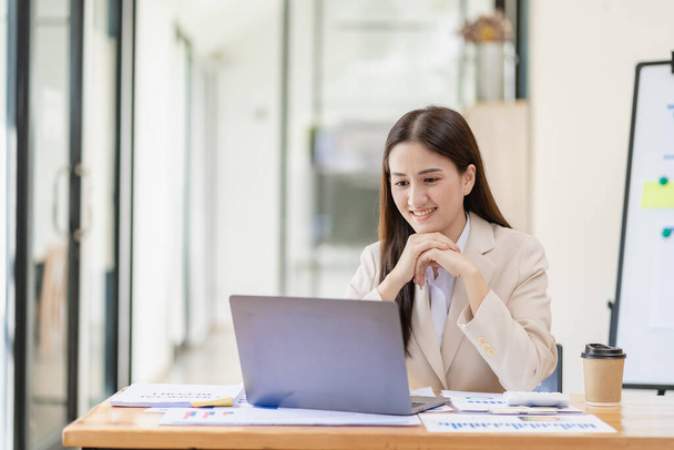 Asiatische Unternehmerin, die im Homeoffice im Finanzbereich arbeitet und Finanzgrafiken auf Dokumenten analysiert, mit Laptop auf dem Tisch im Büro - Foto, Bild